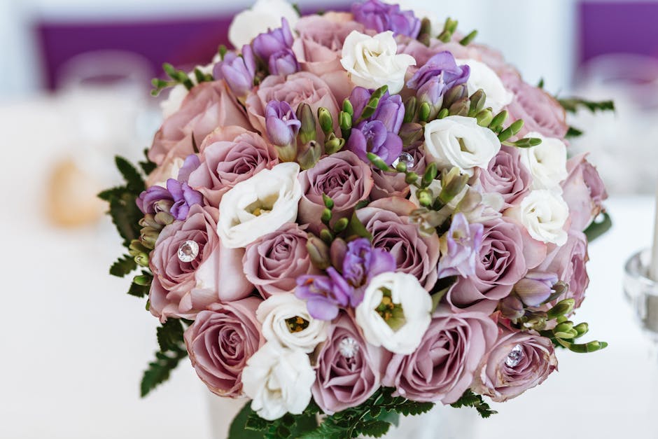 A close-up of a floral bouquet arranged with soft pink roses, white lisianthus, and purple freesias, accented with small green foliage and decorative rhinestones. The bouquet is tightly packed with fr
