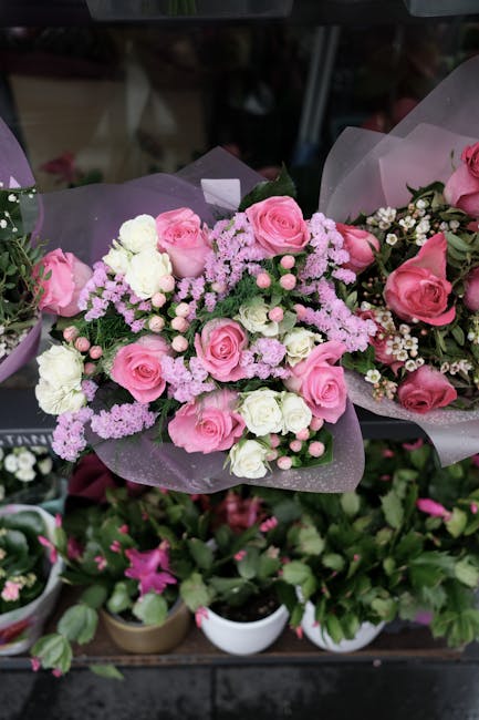 A bouquet of fresh pink roses, white carnations, and delicate light purple statice flowers, arranged with green foliage and wrapped in soft lilac tissue paper. The bouquet is placed on a dark surface,