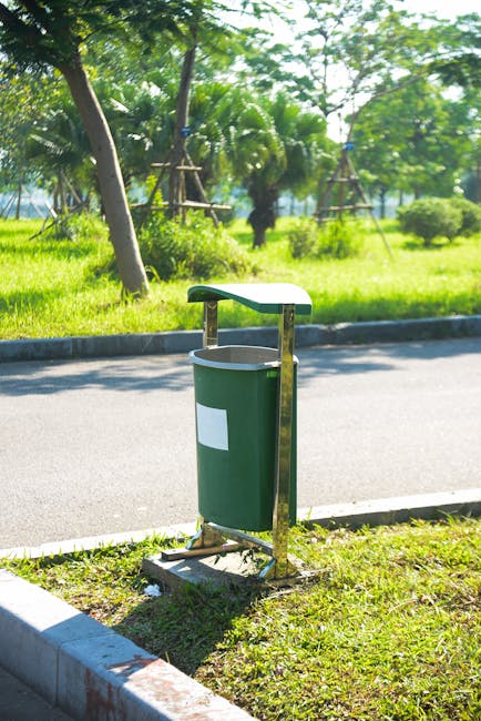 An outdoor scene featuring a green flower or plant waste bin with a white label, positioned on a grassy verge beside a paved road. Behind the bin, there are lush green trees, including palms, with som