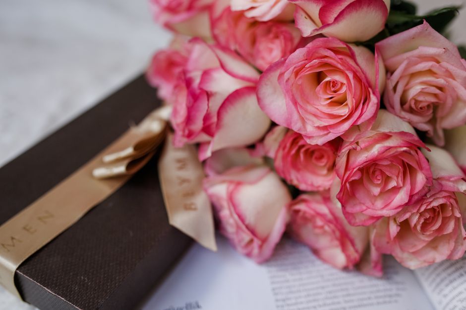 A bouquet of fresh pink and cream roses with delicate petals, arranged in a loose, natural style and placed on a white surface. The roses are partially wrapped in a transparent cellophane, with some s