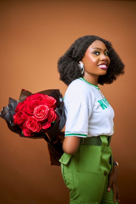 A woman with dark, textured curly hair is holding a large bouquet of fresh red roses wrapped in black paper, positioned behind her. She is wearing a casual white T-shirt with green accents around the