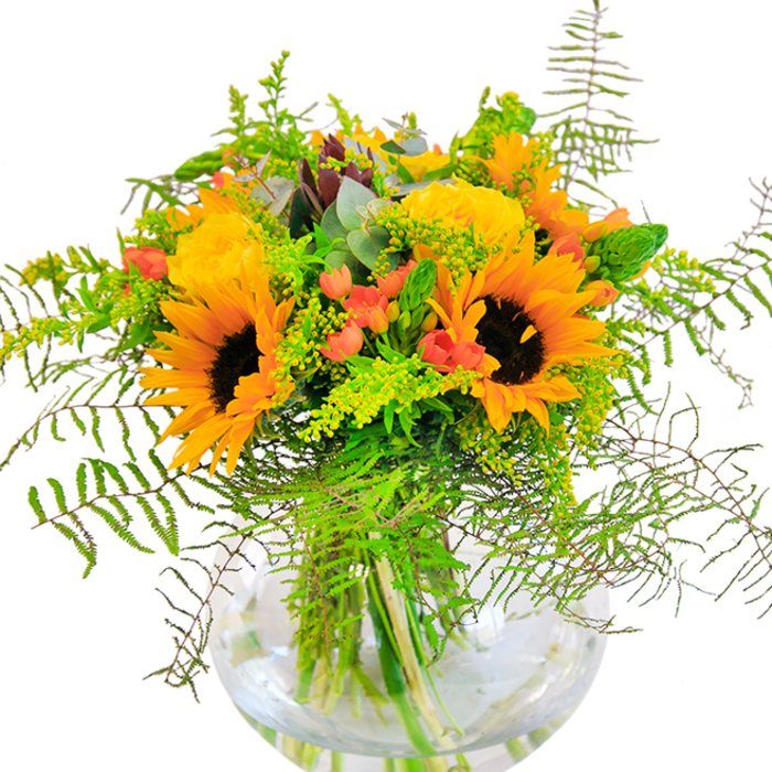 This sunflower and rose bouquet is a lively tribute to the vibrant gardens and close-knit streets of Whetstone. Sunflowers take pride of place, their golden crowns and chocolate centers evoking images of sunny days spent in Friary Park, petals reaching outward in a joyful display. The arrangement is thoughtfully accented by clusters of soft yellow garden roses, each one with a spiral of luxurious petals that imparts a touch of classic sophistication. In between, gleaming red Hypericum berries provide a flash of color, their glossy surfaces catching the eye and introducing a playful brightness. Slender stalks of goldenrod and sprigs of feathery fern cascade delicately among the blooms, their green hues and gentle motion reminiscent of the windswept greenery along Whetstone's tree-lined pathways. Interspersed throughout are hints of blue-green eucalyptus, their subtle shade cooling the palette and evoking the freshness of open fields in late summer. The round glass vase, clear as a drop of dew, anchors the bouquet and gives a glimpse of the tangle of green stems united beneath the surface, their vibrancy magnified in the sunlight that streams through High Road's shop windows. The bouquet's scent is soft yet evocative, a reminder of meandering market mornings and new beginnings-a fitting centrepiece for celebrations, anniversaries, or simply spreading a bit of Whetstone's sunshine at home.