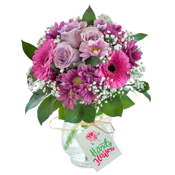 Centered in a crystal-clear, textured glass vase, this exquisitely arranged mixed bouquet showcases the skills of Whetstone's dedicated florists. The arrangement bursts with a carefully balanced palette of lavender, fuchsia, and deep magenta-a modern classic designed to brighten local homes from Woodside Park to Totteridge Lane. The arrangement's heart is made up of several lavender-hued roses, their soft, unfolding petals suggesting a subtly sweet fragrance. Bold fuchsia Gerbera daisies stand out with their large, round petals and striking dark centres, surrounded by clusters of smaller, richly coloured chrysanthemums whose plum and magenta shades offer contrasting texture. Sprigs of dainty white baby's breath are artistically distributed, creating an airy, ethereal feel, while dark green foliage frames the vivid blooms for a natural finish. A rustic strand of raffia ties gently around the vase, holding a simple white tag ready for a heartfelt message. The pristine white background spotlights each nuance, as though this bouquet awaits in a fresh Whetstone flower shop ready for same-day delivery. Whether displayed on a kitchen table overlooking Whetstone Green or presented as a cheerful gift for a birthday or anniversary, this floral arrangement embodies both local artistry and the enduring beauty of thoughtfully chosen flowers.