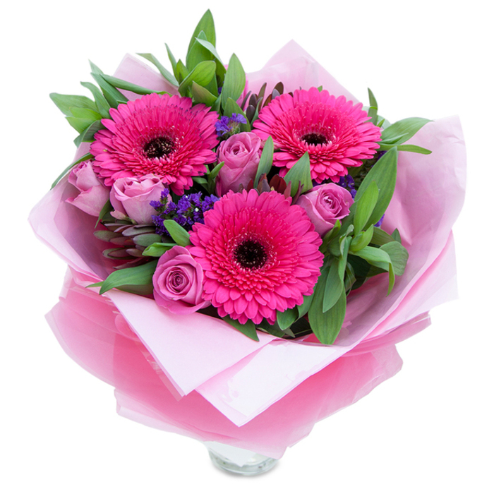 I see a bouquet that balances playful colour with gentle refinement: three rich fuchsia Gerbera daisies open boldly at the centre, each petal catching light and directing attention toward the dark, almost black centres that supply graphic contrast. Nestled among them are roses in a muted lavender-pink-soft, dusty-rose tones where the petals curl inward like tender hands-bringing softness and quiet romance to the arrangement. Scattered clusters of tiny purple filler flowers, likely statice, create pockets of cool violet that lift the palette and add a meadow-like texture between the larger blossoms. A backdrop of generous green foliage, appearing as salal or eucalyptus leaves, gives the bouquet fullness and a crisp, botanical freshness that complements the floral hues. The wrapping reads like a gentle promise: an inner layer of matte pale pink tissue cushions the stems, while a translucent, slightly crinkled fuchsia-pink cellophane wraps the outer form with a light shimmer; thoughtful folds expose the top rim of a clear glass vase beneath, suggesting the bouquet is ready to sit on a windowsill or dining table. The overall impression is one of celebration and comfort-the scent light and floral, the textures velvety and fresh-and it's the kind of arrangement a florist from Whetstone would prepare for a neighbour heading to a small dinner party, a birthday visit, or simply to lift the mood in a nearby Barnet home.