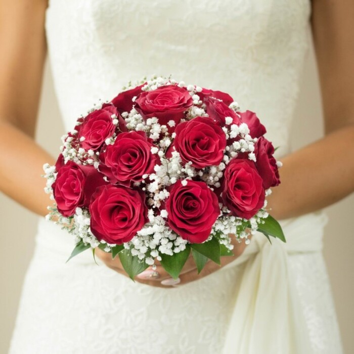 Captured in a tender mid-torso view, the image showcases a bride in Whetstone clutching a classic round bouquet-a beloved emblem of romance designed for North London ceremonies. The central focus is the vibrant cluster of deep red roses, their soft, velvety petals unfurling in rich, undulating layers, each bloom hinting at devotion and grace. These dominant roses, beautifully arranged for maximum fullness, are embellished by a multitude of airy white gypsophila blossoms. The baby's breath, with its cloudlike clusters, weaves throughout the roses and contributes a refined, dreamy contrast that feels almost ethereal-a gentle nod to Whetstone's tranquil gardens and green spaces. Just below the floral display, fresh green leaves offer a burst of natural color, accenting the hand-tied bouquet while anchoring it in the visual landscape of North London. The bride's manicured hands gently support the base, conveying the care taken by a skilled Whetstone florist. Her wedding gown, crafted in lace or delicately embroidered fabric, adds rich texture and sophistication, with a flowing sash trailing down her side. A softly blurred, neutral-hued background ensures each intricate detail of the bouquet and dress is bathed in flattering, gentle light-much as one might find on a calm Whetstone afternoon. The composition evokes the traditions and heartfelt joy surrounding local weddings, providing inspiration for anyone seeking an arrangement infused with elegance and enduring romance.