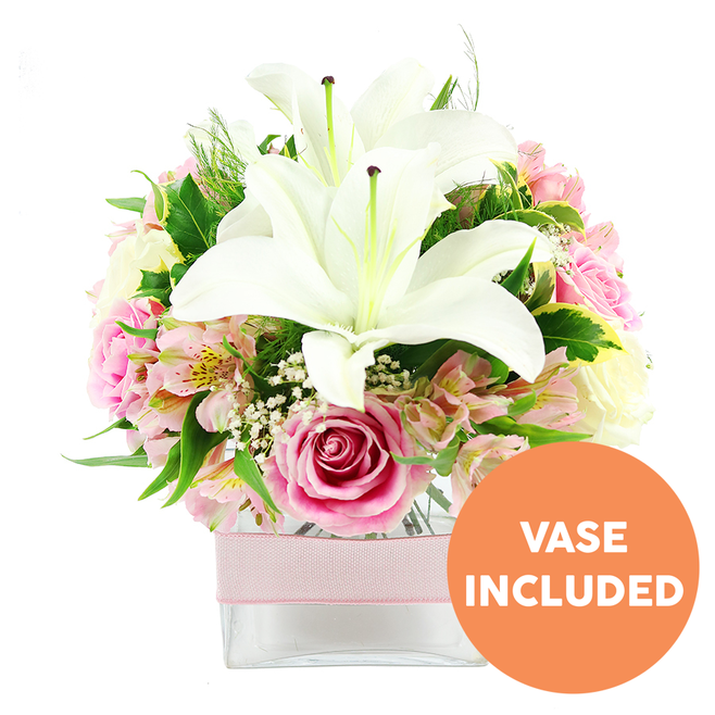 A refined, dome-shaped bouquet of white lilies and pink roses is displayed against a bright white background, the colours standing out with crisp clarity as though captured in the soft midday light over Whetstone. Two grand white Oriental lilies form the crown of the arrangement, their petals wide and slightly recurved, with rich reddish-brown stamens poised at the centre like tiny sculptures. Encircling these feature blooms is a generous ring of pink roses, their shades ranging from delicate blush to medium rose pink, each flower at a different stage of opening. Some roses remain closed in neat, rounded buds, while others have fully unfurled into open, layered rosettes, the petal edges gently curled and velvety to the eye. Nestled between them are clusters of alstroemeria, their slender petals washed in pale pinks and creamy whites, each blossom marked with fine streaks of yellow and soft freckles that add a sense of intricate craftsmanship. Wisps of white baby's breath, with dozens of tiny star-shaped blooms, drift between the larger flowers, creating a hazy, romantic halo. Around the base, a collar of mixed greenery-broad, shiny leaves, subtle variegation, and airy, fern-like fronds-adds fullness and depth, echoing the lushness of gardens tucked away on the residential streets around Whetstone and Totteridge. The entire composition is nestled neatly into a low, square glass vase, the clear sides revealing straight green stems submerged in water, and a pale pink ribbon, smooth and satin-finished, is wrapped horizontally around the vase for a polished, gift-ready look. In the bottom right corner, a vivid orange circular badge carries the words VASE INCLUDED in bold white capitals, clearly indicating that the glass container is part of the floral gift. The design feels like a ready-to-place centrepiece for a Whetstone home, perfect for birthdays, anniversaries, or a thoughtful hospital visit at nearby Barnet Hospital, offering beauty and comfort in equal measure.